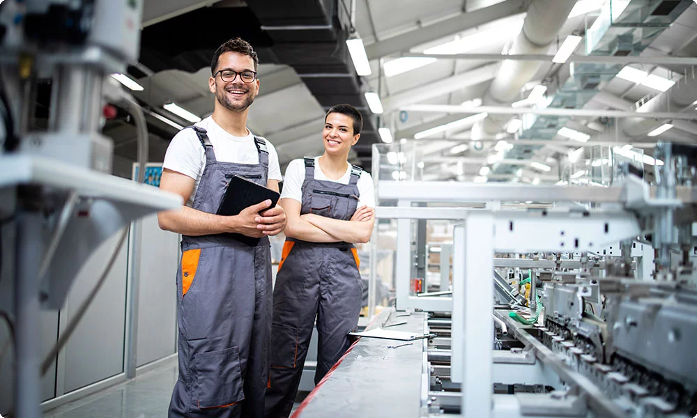 Zwei Mitarbeitende in Arbeitskleidung stehen lächelnd in einer modernen Werkhalle neben einer Produktionsanlage – das Bild unterstreicht die Bedeutung von Gehörschutz am Arbeitsplatz zur Vermeidung von Hörschäden in lärmintensiven Umgebungen.